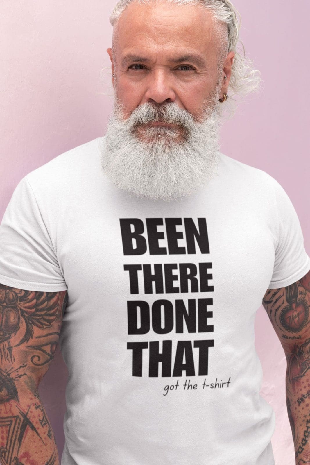 A bearded man with white hair and tattooed arms stands against a light pink background, confidently wearing the Been There, Done That T-Shirt and embodying the bold attitude of this statement piece.
