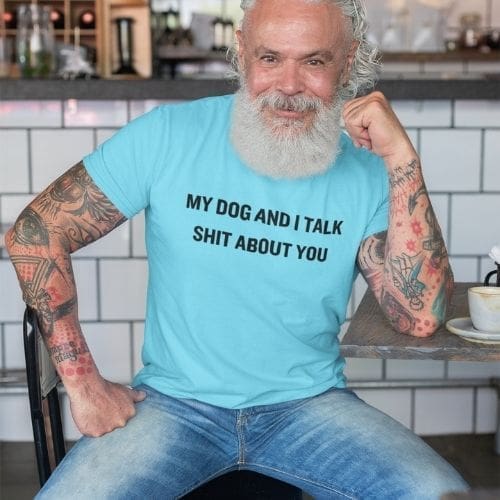 A smiling older man with tattooed arms relaxes in a cafe, wearing the My Dog and I Talk Shit About You T-Shirt, enjoying coffee as he sits casually at the table.