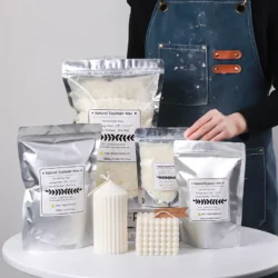 A person in a blue apron stands behind several silver bags of Natural Soy Wax For DIY Handmade Candles, alongside two handmade candles””one tall and ribbed, the other shaped like a bubble cube””displayed on a white surface.