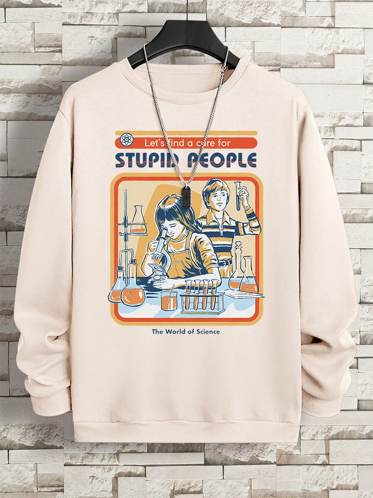 A beige Lets Find A Cure For Stupid People Sweatshirt hangs on a hanger against a brick wall. Its graphic shows two people in a science lab with the text “Let”™s find a cure for STUPID PEOPLE” and “The World of Science.” A silver chain necklace is draped over it.