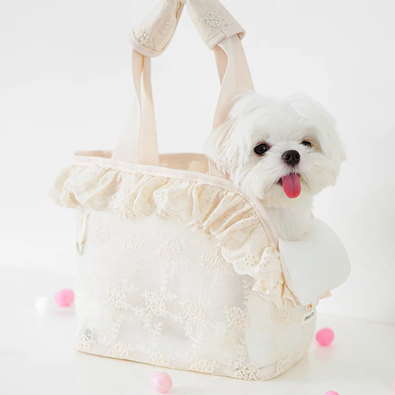 A small white dog with fluffy fur sits inside a cream-colored Cute Lace Carrier Bag for Dogs with ruffled edges, tongue out, surrounded by pink and white pom-poms””showcasing one of the cutest pet carrier bags.