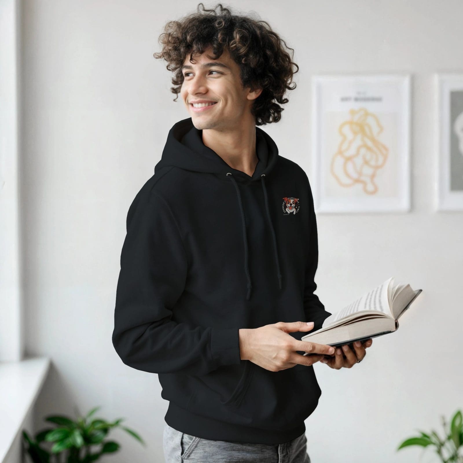 A young man with curly hair stands indoors near a window, smiling and looking to his left while holding an open book. He wears The Exclusive Spencer Vibes Hoodie, which features a small tiger graphic. Framed art and green plants are in the background.