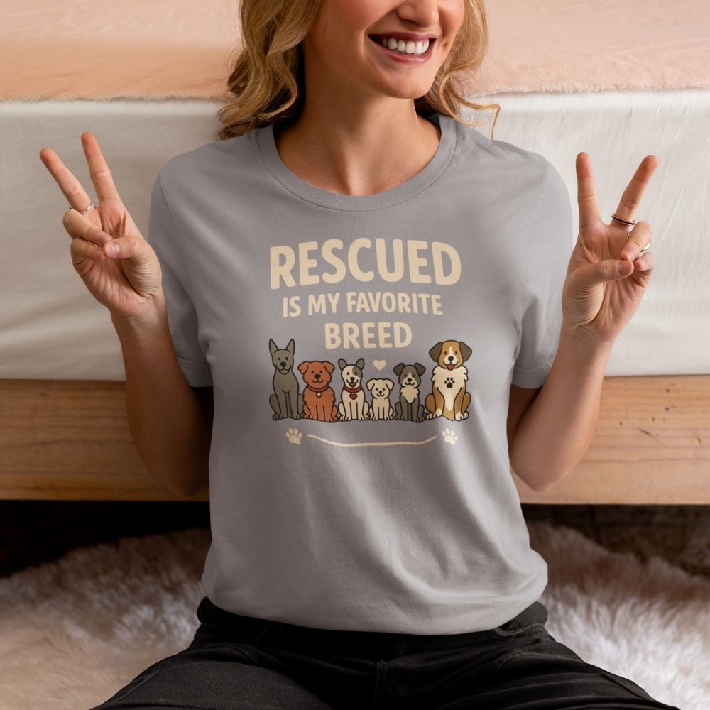 A woman sits cross-legged on a soft carpet, flashing peace signs and wearing the Rescued is my favourite breed gray T-shirt featuring six dogs. Part of a bed is visible in the cozy setting.