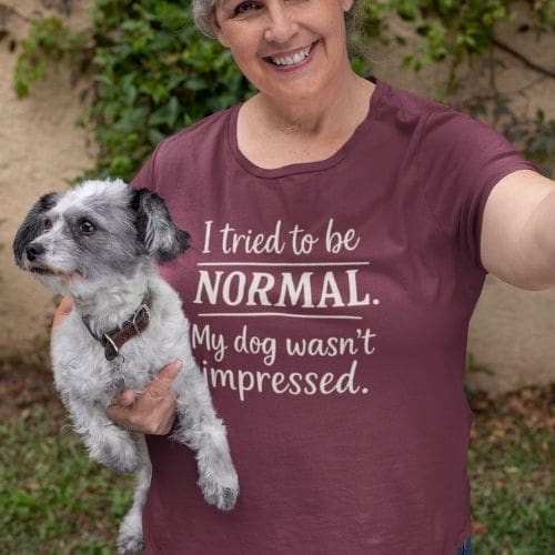 A smiling woman with short gray hair holds a small black-and-white dog, wearing a maroon Heavyweight Unisex Crewneck T-shirt | GildanÂ® 5000 that reads, I tried to be NORMAL. My dog wasn”™t impressed. Green foliage and a tan wall are behind her.