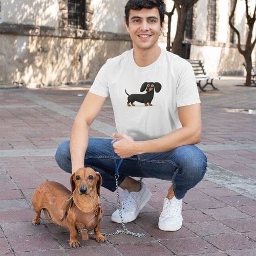 A smiling man wearing a Heavyweight Unisex Crewneck T-shirt | GildanÂ® 5000 with a cartoon dachshund design kneels outdoors on pavement, holding a brown dachshund on a blue leash. He wears jeans, white sneakers, and has short dark hair; trees and benches behind.