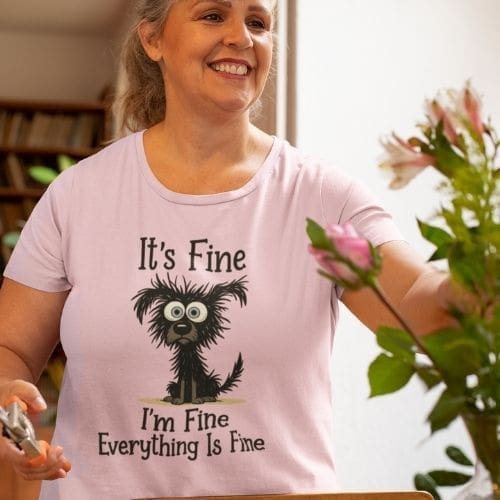 A woman arranges flowers indoors, wearing a light pink Heavyweight Unisex Crewneck T-shirt | GildanÂ® 5000 featuring a cartoon dog and the phrase It”™s Fine. I”™m Fine. Everything Is Fine. Bookshelves and blurred flowers are in the background.