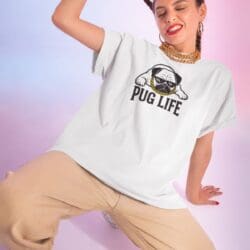 A woman in tan pants and a white Pug Life T-shirt dances joyfully. She wears gold jewelry, red lipstick, has her hair tied back, and stands against a soft pink and purple gradient background.