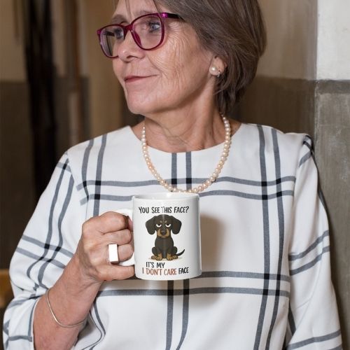 An older woman with short gray hair, red glasses, and a pearl necklace smiles slightly as she holds the I Dont Care Face Mug, featuring a cartoon dog. She wears a white blouse with a black plaid pattern and looks to the side.