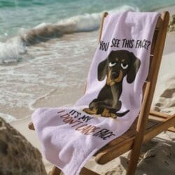 A wooden beach chair rests on sand by gentle waves, draped with the Funny Dog Beach Towel””Perfect for Pet Lovers””featuring a cartoon dachshund and the saying “YOU SEE THIS FACE? ITS MY I DONT CARE FACE” in bold black and pink letters.