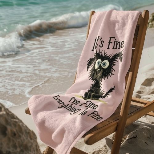 A light pink towel with a cartoon of a frazzled black bird and the text “It”™s Fine. I”™m Fine. Everything Is Fine” drapes over a wooden chair on a sandy beach near gentle waves under sunlight.