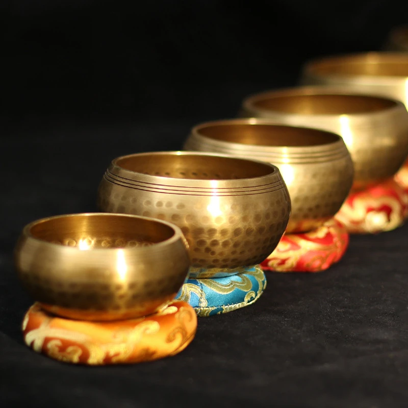 Five Nepal Hand-made Buddha Sound Bowl | Brass Meditation Singing Bowls of different sizes are arranged diagonally on vibrant embroidered cushions, their hammered brass surfaces gleaming against a dark background.