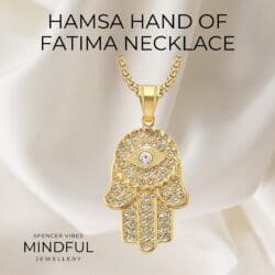 A gold Turkish Evil Eye Hamsa Hand of Fatima Pendant Necklace, made from stainless steel and iced out with crystals, hangs on a gold chain for women and men. Displayed on silky white fabric beneath the text "HAMSA HAND OF FATIMA NECKLACE.