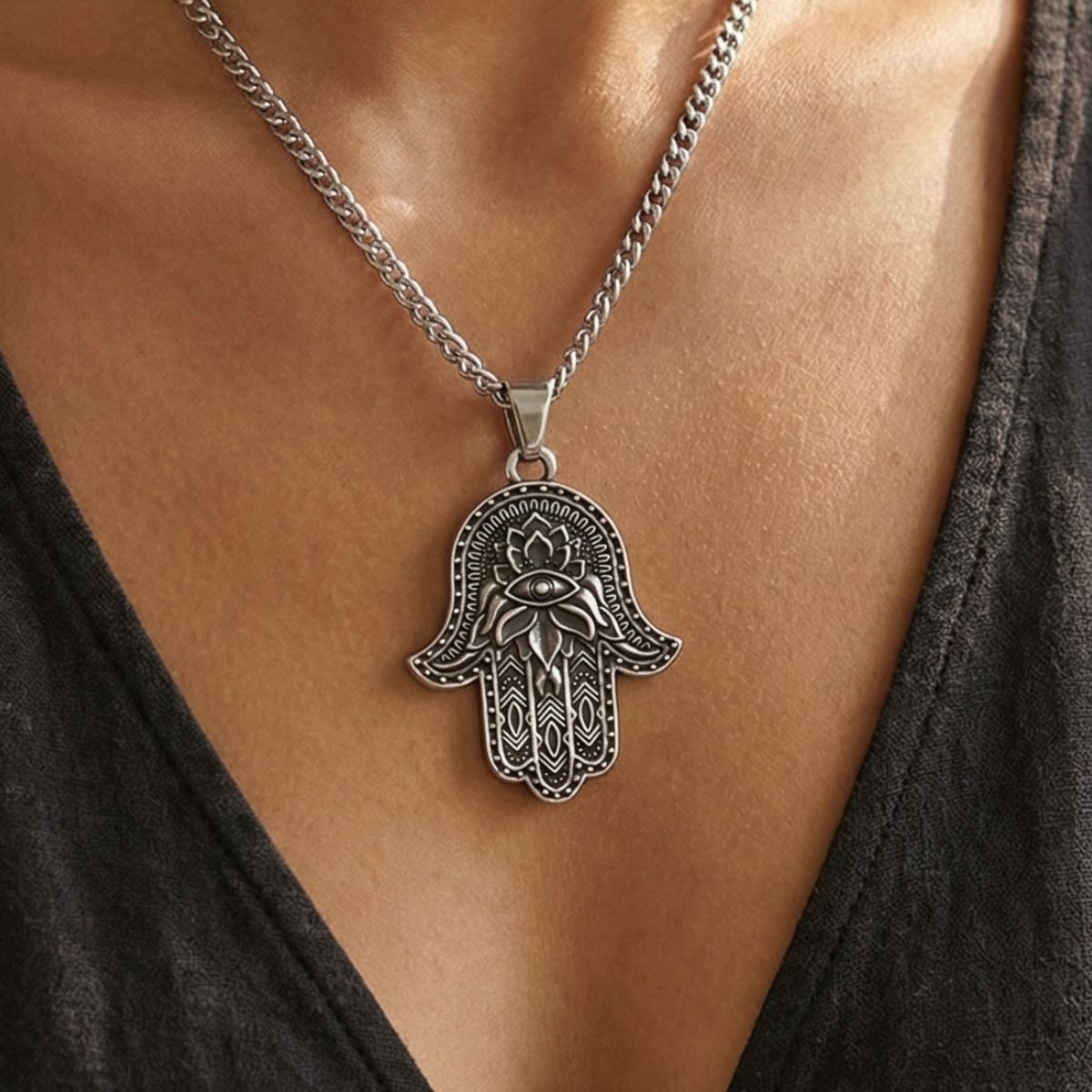 A person wears the Hamsa Hand of Fatima Pendant, a silver chain necklace featuring intricate patterns and a central eye design. Their skin and part of a black V-neck top are visible in the background.