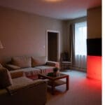 A cozy living room with beige walls, two tan sofas with patterned throws, a wooden coffee table with a plant, and a wall-mounted TV glowing with red light, perfect for an evening routine in a warm atmosphere.