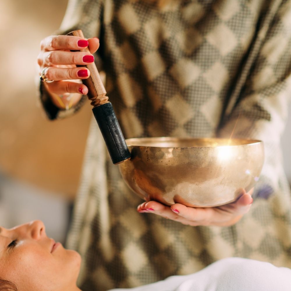 A person holds a brass singing bowl and gently strikes it with a wooden mallet. The calm lifestyle scene features another person lying down, eyes closed, possibly receiving a sound therapy session focused on peace and relaxation.