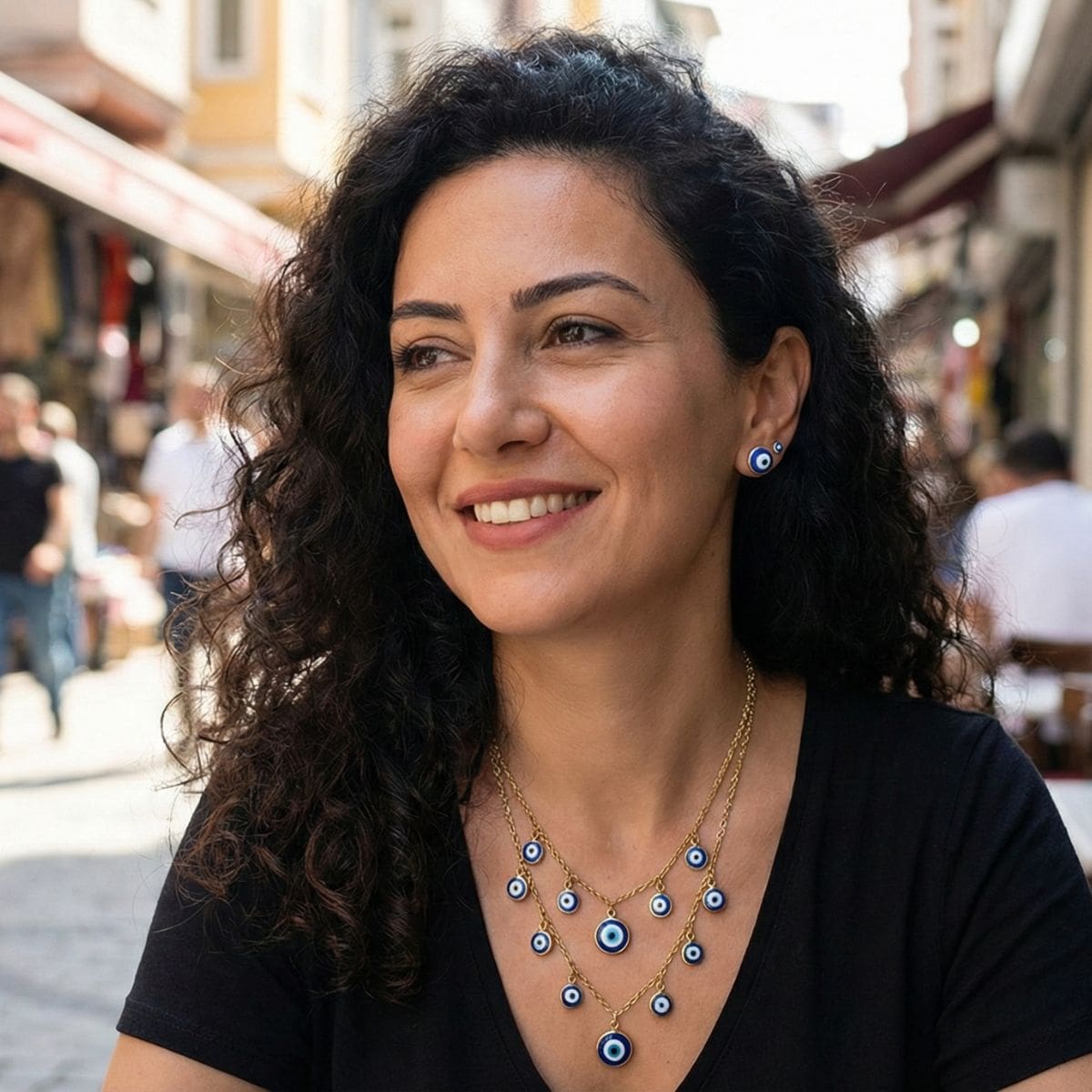A woman with curly dark hair and a black top smiles while sitting outdoors at a busy street café. She wears matching blue evil eye symbolic jewellery, including a sterling silver pendant. The background is slightly blurred, showing people and shops.