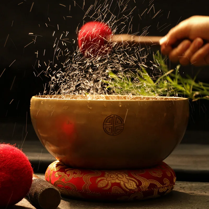 A hand strikes the Nepal Handmade Full Moon Singing Bowl with a red mallet, sending water droplets upward. The bowl sits on a patterned red cushion, filled with grass-like plants and displaying an intricate circular symbol in the background.