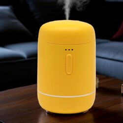 A yellow Essential Oil Waterless Nebuliser with a central button and indicator lights sits on a wooden table, releasing a visible mist. A dark sofa and cushions are softly blurred in the background.