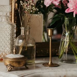 A marble countertop displays a brass candlestick, a decorative brass box with a lid, hints of aromatherapy for sleep, a cut-glass decanter, and two glass vases with pink flowers and delicate greenery—all reflected in a large ornate mirror.