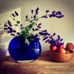 A round blue glass vase with purple flowers and green stems sits on a wooden table beside a bowl of apples, creating a calming scene perfect for aromatherapy for anxiety, against an off-white wall with soft natural light and gentle shadows.
