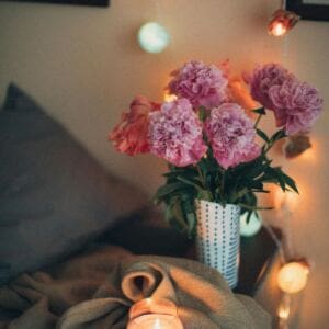 A vase with pink peonies sits on a cozy, softly lit surface next to aromatherapy oils and a lit candle. Warm string lights in the background cast a gentle glow, while a blanket and pillows enhance the inviting, relaxed atmosphere.