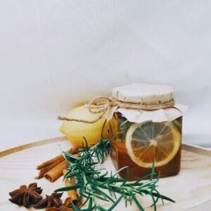 A jar of honey with lemon slices inside, covered with paper and twine, sits on a wooden tray. Aromatherapy elements like cinnamon sticks, star anise, and fresh rosemary surround it, along with a whole lemon, against a plain white background.