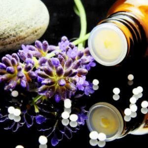 A close-up of a purple lavender flower beside an open amber glass bottle labeled "oils not to diffuse," with white round pills scattered on a shiny black surface. A smooth gray stone sits in the background, reflected in the glossy scene.