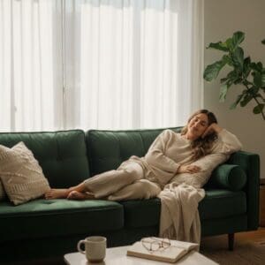 A woman in cozy beige clothes relaxes on a green sofa, eyes closed and smiling gently. She leans on a pillow and blanket, with a large plant nearby. Soft daylight streams in, while her CES devices UK rest beside her coffee mug and book.