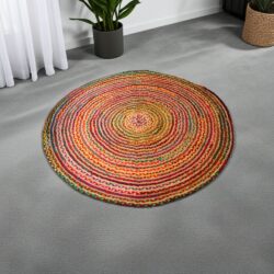 The Round Jute and Recycled Cotton Rug - 150 cm, featuring vibrant concentric rings, lies on a light gray carpet. Nearby, a potted plant and woven basket accent the white wall and sheer curtains in the background.