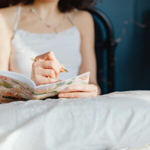 A woman in a white camisole sits on a bed, journaling in a floral-patterned notebook and reflecting on manifesting mistakes. She is partially visible from the shoulders down, with soft natural light highlighting the pillow and bedspread in the foreground.