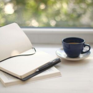 An open notebook with lined pages lies atop a closed book, black pen ready for manifesting money. Beside them, a dark blue cup of coffee sits on a matching saucer on a white table by a window with blurred greenery outside.