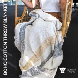 A person sits in a woven chair with the Boho Cotton Throw Blanket – Mellow Mattala Neutral Stripe Throw draped over their lap, featuring cream with wide mustard yellow and gray stripes. “BOHO COTTON THROW BLANKET” appears on the left; Spencer Vibes logo at bottom right.