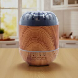 The Nordic Minimalist Ultrasonic Aroma Diffuser – Wood Effect, with a rounded wood-grain design and grey vented top with timer buttons, sits on a light wooden table in a bright, sunlit kitchen with blurred white cupboards.