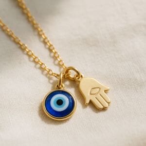 A gold chain necklace with two pendants: a round blue charm symbolizing nazar evil eye meaning and a gold Hamsa hand charm. Both rest on a soft beige fabric, with the Hamsa featuring a simple engraved eye design in the center.