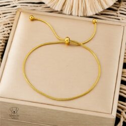 The Adjustable Gold Slider Bracelet – Minimal Everyday Jewellery, featuring round end beads, sits on a light beige jewelry box with the Spencer Vibes logo on the corner. The background shows a woven surface and decorative accents.