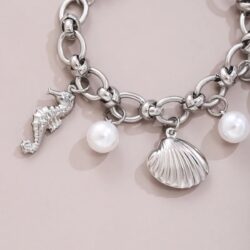 The Silver-Tone Ocean Charm Bracelet with Pearl Details features large chain links and dangling charms—a seahorse, a seashell, and two round pearls—displayed on a light beige surface.