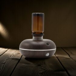 The Intelligent Mini | Motion-Activated Waterless Nebuliser features a wood grain base, translucent amber glass top, and a glowing white ring at its USB-C powered base, all set on a wooden surface beneath soft ambient lighting.