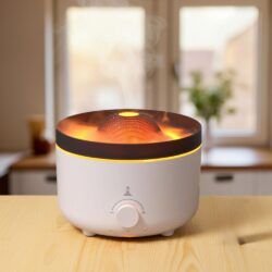 The Grand Volcano Sleep Kit’s white 560ml nebuliser emits mist upward from a wooden table, its top glowing with a warm orange flame-like light. Blurred windows and a potted plant accent the bright background. Lavender bundle included.