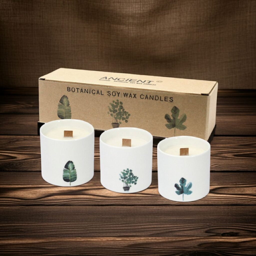 Three white Large Botanical Candles – Victorian Peony, each with wooden wicks and unique botanical illustrations, are displayed in front of a brown box labelled BOTANICAL SOY WAX CANDLES on a wooden surface with a fabric backdrop.