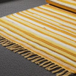 The Large Rag Rug – 150 × 90 cm (Sunrise Orange) displays horizontal yellow, orange, and white stripes with fringed tassels on the edge, resting on a gray carpeted floor and one corner rolled up to show more of its pattern.