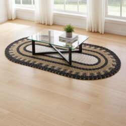 An Oval Seagrass Rug Black, White & Tan Classic (60 × 120cm) anchors the room beneath a glass-top coffee table with black metal legs, topped with books and a potted plant. Light wood floors and white-curtained windows enhance the minimalist space.