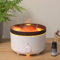 A white essential oil diffuser emits orange-lit mist resembling fire. It sits on a wooden surface near a potted fern and a small bottle labeled “Lavender Essential Oil.” Warm light glows from inside the diffuser, creating a cozy atmosphere.