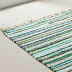 The Large Rag Rug - 150x90cm, premium quality in Natural Greens, with green, blue, white, and grey stripes plus fringe lies on a beige carpet in a bright room, adding a colourful accent to the neutral decor.