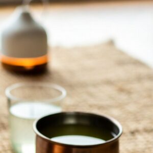 A close-up of a metal cup filled with green tea sits on a woven surface. Behind it, a glass of water and a ceramic diffuser glow softly, creating a calm atmosphere—perfect for moments of balance and mindful Ayurvedic Wellness.
