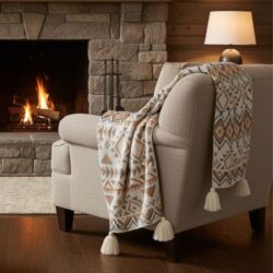 A beige armchair with a Boho Geometric Throw (150 × 220 cm) draped over the back sits on a wooden floor by a stone fireplace with a lit fire and a softly glowing table lamp, creating a warm, rustic atmosphere.