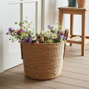 The Hand-Braided Seagrass Sanctuary Basket – Natural Woven Storage sits on a wooden floor, filled with garden tools and wildflowers. Nearby, a wooden stool and potted plant rest by a sunlit window in the background.