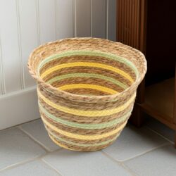 A set of Seagrass & Straw Vase-Shaped Baskets in yellow and green sits empty on a light grey tiled floor near a white panelled wall and brown wooden cupboard. Handmade from natural fibres with alternating stripes, these baskets come as a set of three.