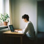 A young person with short dark hair sits at a wooden table, typing on a laptop near a window with natural light—embracing mindfulness for remote workers. A potted plant adds calm, while the room’s cream walls and minimal decor create ideal home essentials.