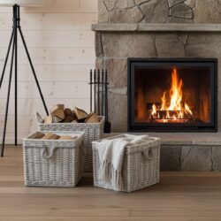 A cosy living room features a stone fireplace, firewood and a grey knitted blanket in the Nesting Wicker Storage Baskets with Liners – Set of 3 on the wooden floor, plus a black fireplace toolset, tall floor lamp, and light wood walls.