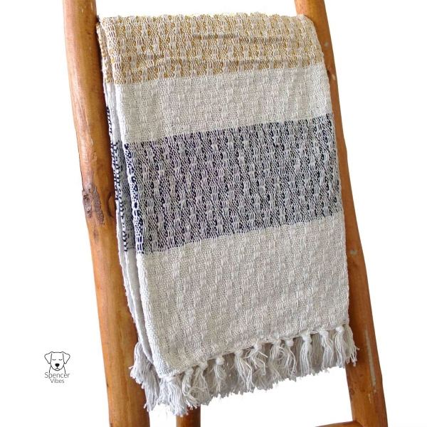 The Boho Cotton Throw Blanket – Mellow Mattala Neutral Stripe Throw, featuring thick beige, black, and off-white stripes, textured patterns, and fringe tassels, drapes over a ladder with a small dog logo and Spencer Vibes in the corner.